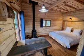 Intérieur chaleureux tout en bois d'un hébergement insolite aux Cabanes du Lac de Pierre-Percée en Meurthe-et-Moselle, révélant une chambre douillette avec un grand lit double, une table rustique et un poêle à bois.