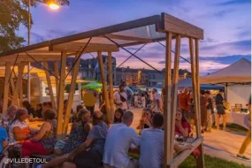 Des personnes attablées chaleureusement sous des structures en bois lors d'une soirée guinguette estivale à Toul en Meurthe-et-Moselle, sous un ciel de coucher de soleil aux nuances roses et violettes.