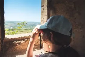 Une jeune fille avec une casquette regarde avec des jumelles à travers l'ouverture en pierre d'un monument, admirant un point de vue panoramique sur la nature en Meurthe-et-Moselle.