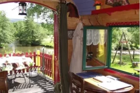 Vue depuis l'intérieur en bois d'un hébergement insolite aux Roulottes de la Meurthe, s'ouvrant sur une terrasse privative avec table et chaises au bord d'une rivière calme en Meurthe-et-Moselle